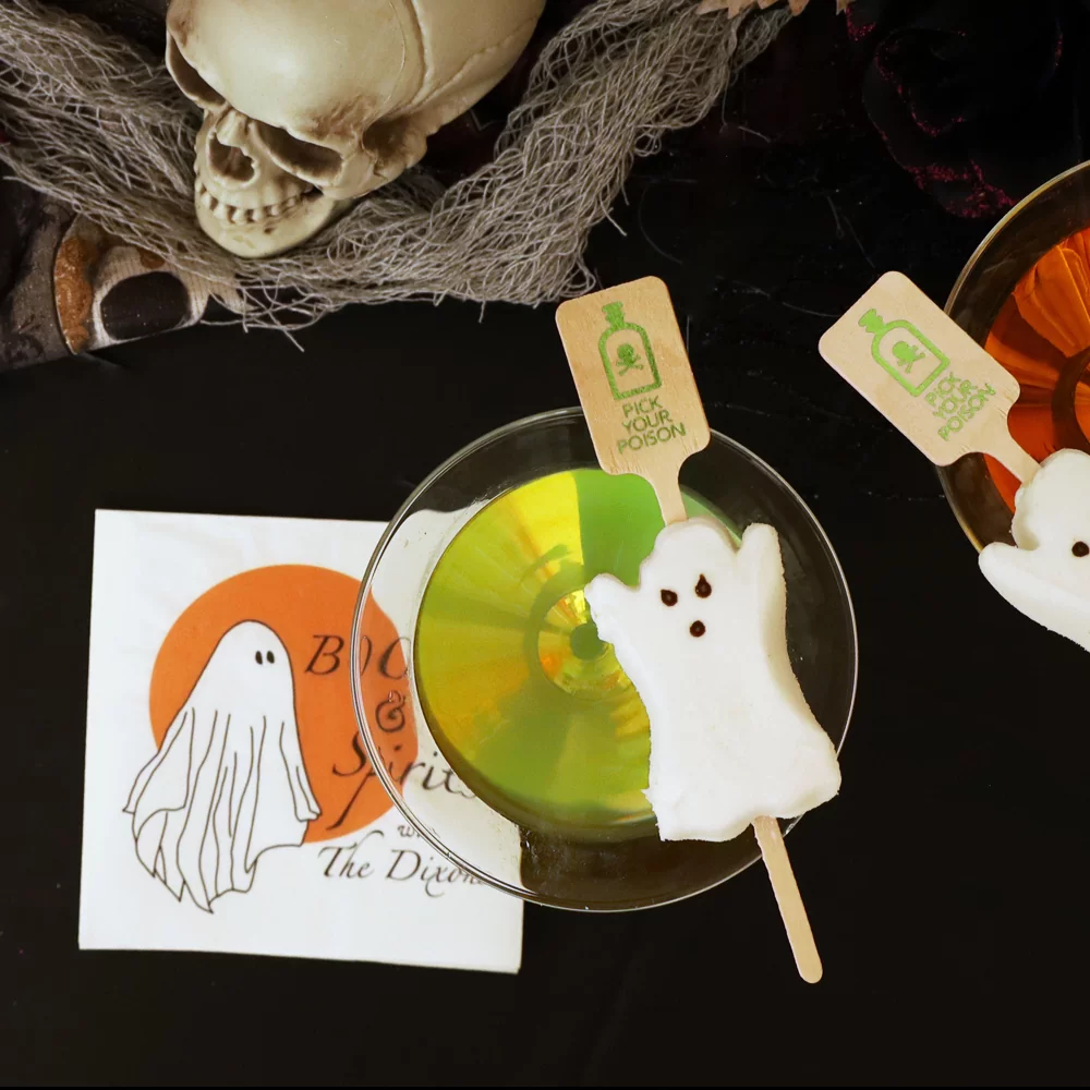 Ghost Marshmallow appetizer on a stir stick place on a martini glass. Cocktail napkin underneath that says "Boos and Spirits"