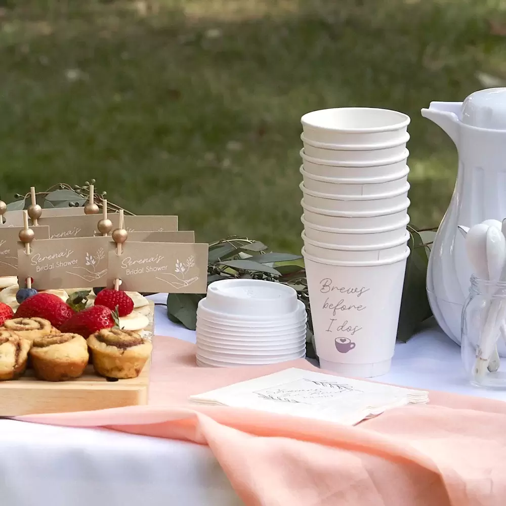 Coffee Cups stacked on a table that say “Brews Before I Do’s” next to some pastries