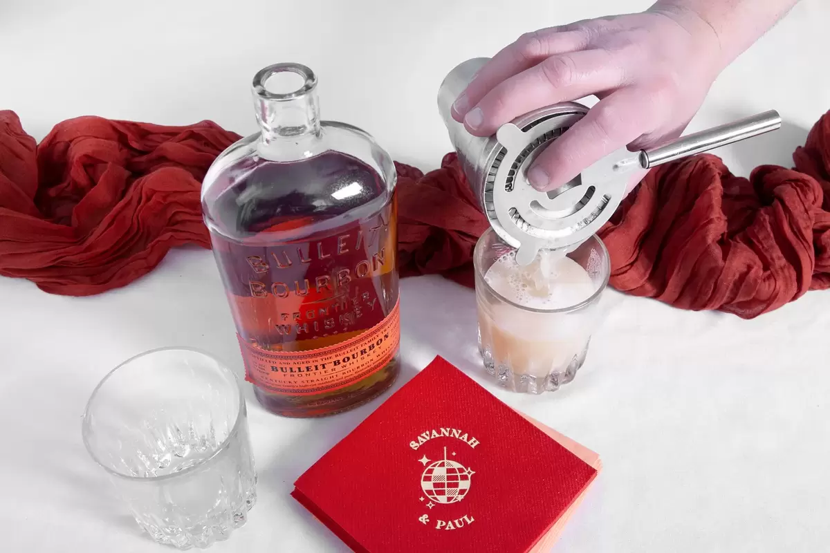 Hand pouring whiskey sour from a cocktail shaker into a glass.