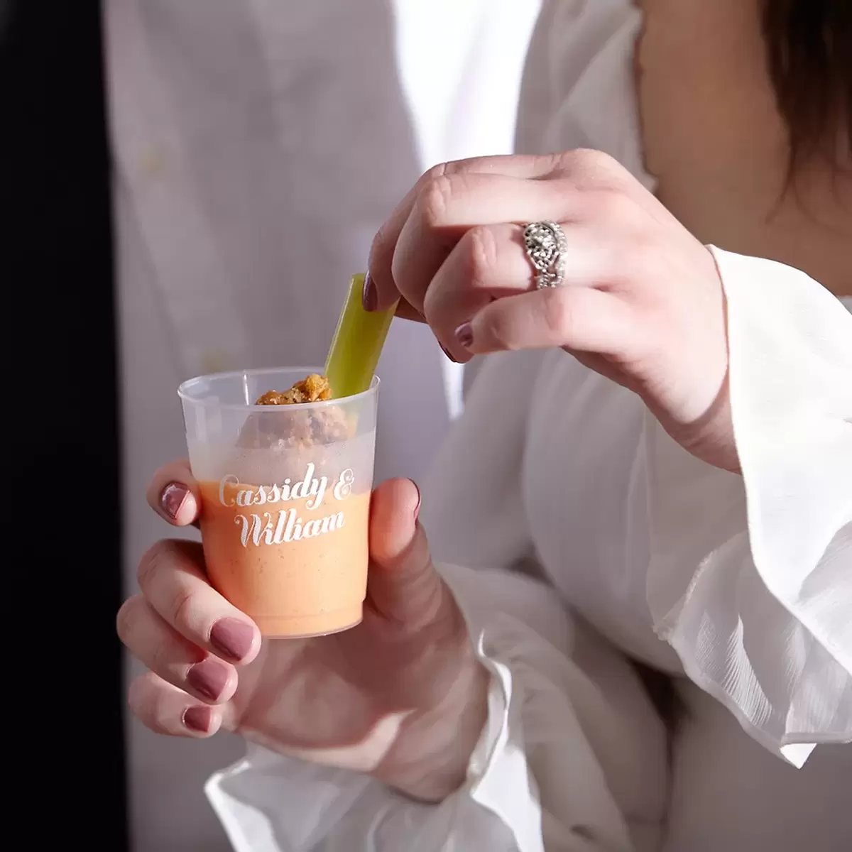 Custom plastic shot glass with 2 names printed on it. The shot glass is filled with sauce and is shown with a hnd dipping a chicken wing into the cup. 